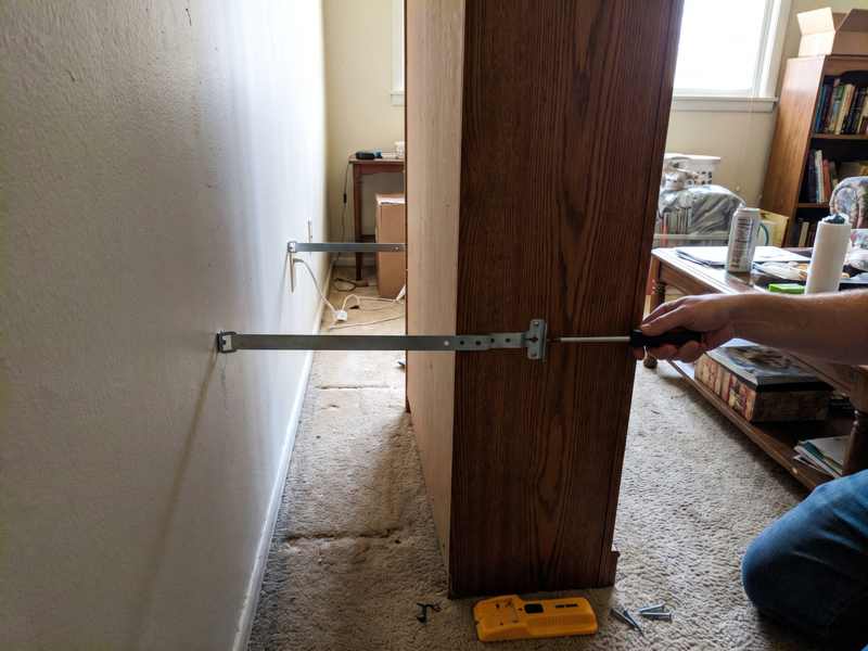 An anti-tip bracket and strap being fitted from the back of a tall bookcase to the wall