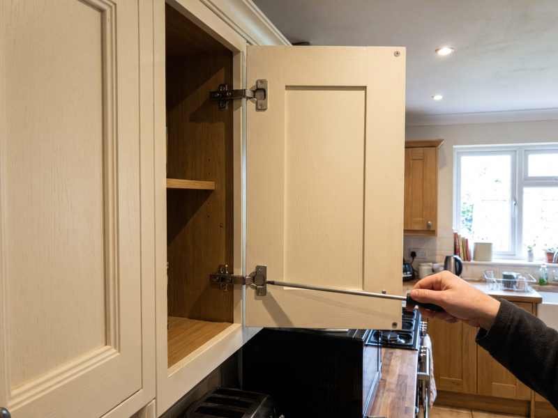 A screwdriver adjusting a concealed hinge inside an open kitchen cabinet door