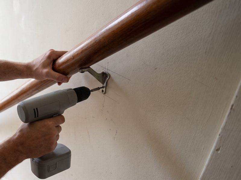 A wooden stair handrail held against a plaster wall while a screw is driven through its bracket