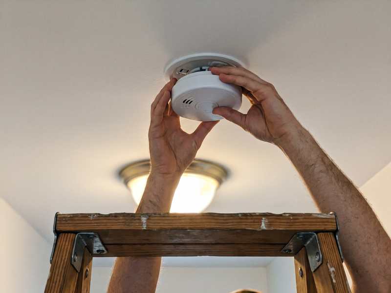 Hands twisting a smoke alarm onto its ceiling mounting plate in a hallway