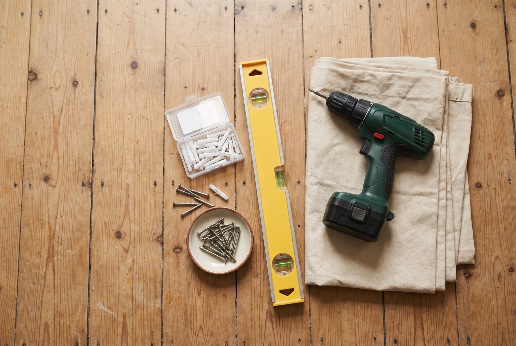 A drill, level, anchors and screws laid out on a wooden floor