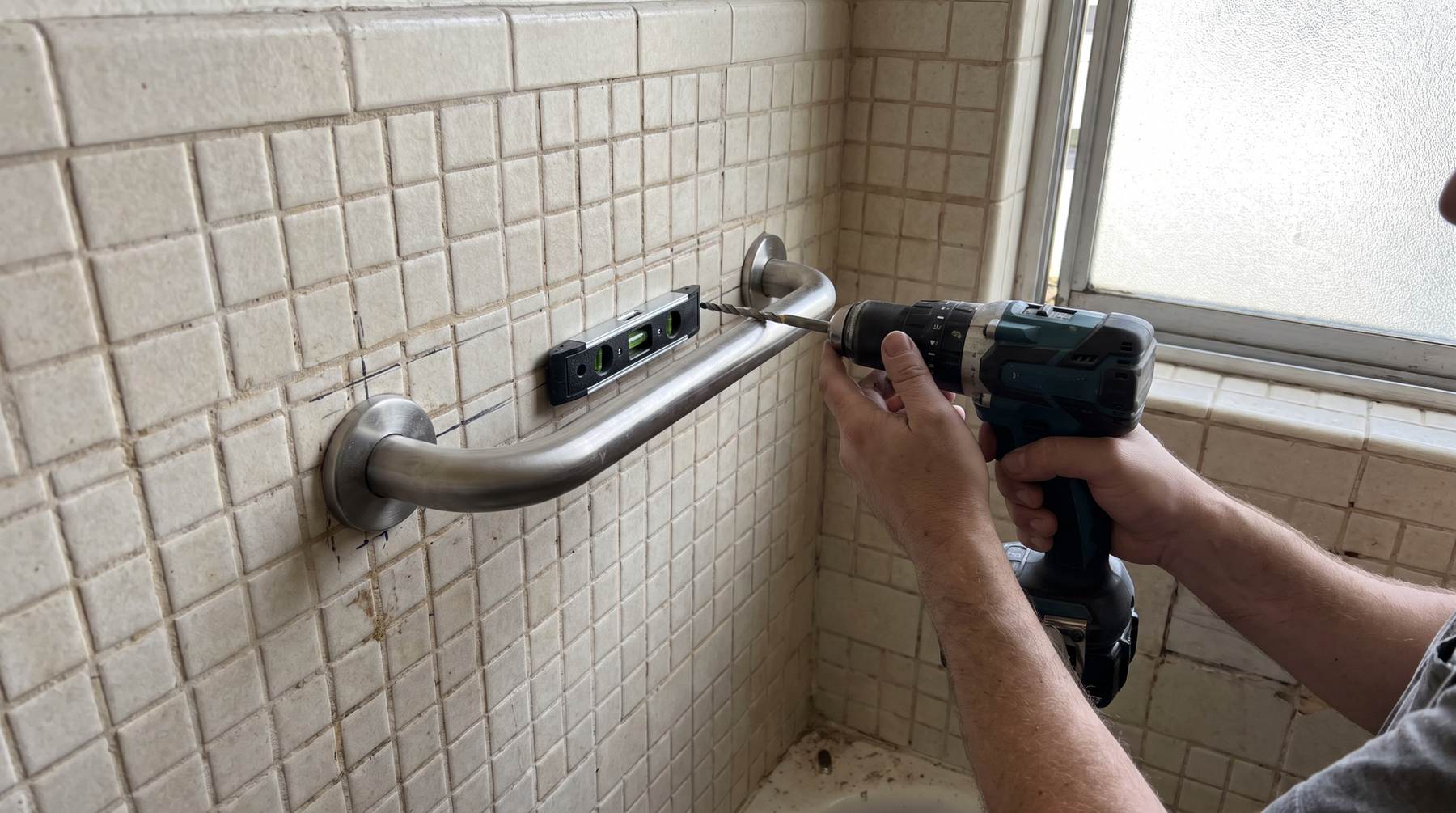 A grab bar held level against a tiled bathtub wall while a fixing is driven through the flange